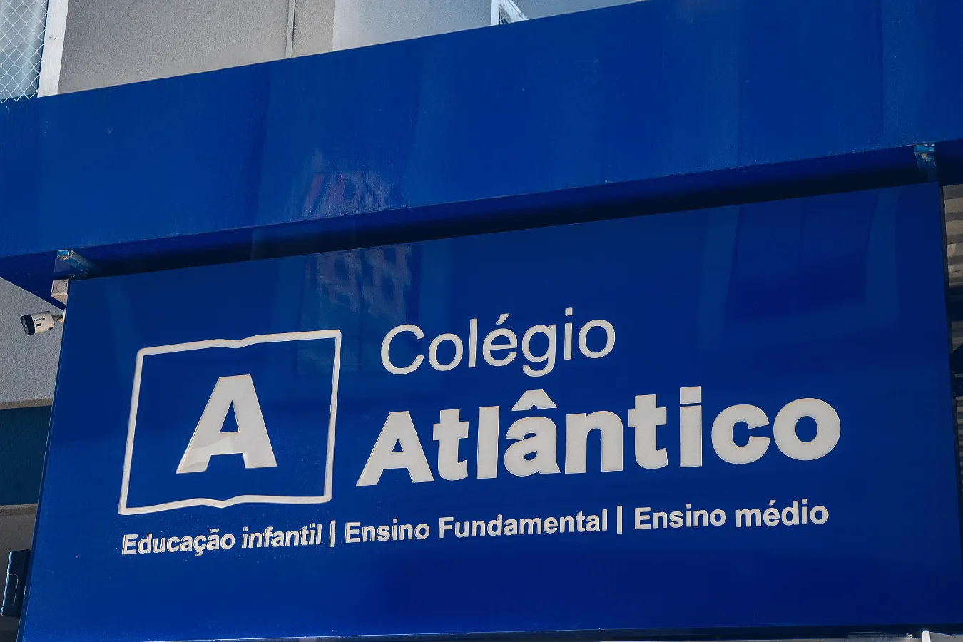 Placa de identificação do Colégio Atlântico com logotipo azul e branco, destacando as etapas de ensino oferecidas: Educação Infantil, Ensino Fundamental e Ensino Médio.